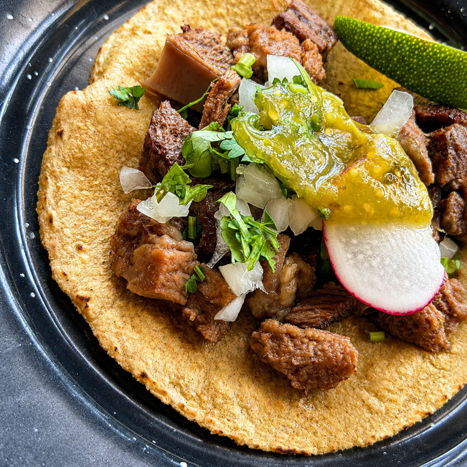 Open tortilla topped with braised meat, melted cheese, avocado, radish, onion, and cilantro on a black plate.