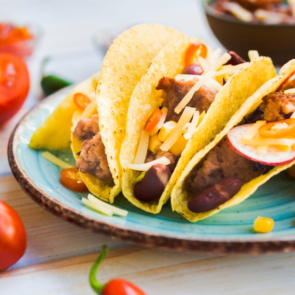 Three crispy tacos filled with grilled meat, onions, peppers, and tomatoes on a turquoise plate.