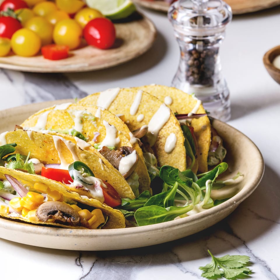 Plate of crispy tacos filled with vegetables, mushrooms, greens, and creamy sauce, with fresh tomatoes and pepper grinder in the background.